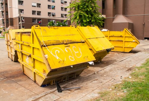 A large yellow metal skip situated outdoors on a paved surface, positioned near a grassy area and in front of a multi-storey residential building with brown and beige exterior finishes. The skip has a rectangular shape with reinforced side panels, visible welding seams, and a rusted finish in some areas, indicating wear and exposure to weather. The front panel features faded white graffiti markings, and the skip is tilted slightly with the front lowered towards the ground. Behind it, there are two additional yellow skips aligned along the pavement, partially obscured by the main skip in the foreground. The environment suggests a typical urban setting for rubbish or waste collection, with some small debris and dirt on the ground nearby. The scene is lit naturally, likely during daytime, highlighting the weathered texture of the metal and the surrounding context of a low-traffic residential area, aligning with services offered by Commercial Waste Uxbridge for commercial rubbish removal.