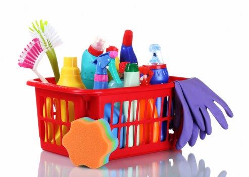 The image shows a red plastic shopping basket filled with various cleaning and household supplies, placed on a white surface. The basket contains multiple brightly coloured spray bottles, brushes, sponges, and gloves, all arranged in a manner typical of cleaning products used for domestic or commercial waste management. To the front of the basket, there is an orange cleaning sponge with a wavy edge, and a pair of purple rubber gloves are draped over the right side of the basket, hanging down slightly. The background is plain white, with no additional environmental details visible. The overall scene suggests an assortment of cleaning equipment commonly associated with rubbish removal and waste disposal tasks, which could be relevant for a professional waste management service such as Commercial Waste Uxbridge serving local businesses in the Uxbridge area.