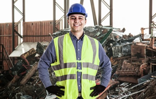 A young male worker wearing a blue hard hat, high-visibility yellow safety vest, and black gloves is standing inside an industrial warehouse or waste processing facility in Uxbridge. Behind him, there is a large pile of mixed construction debris and rubbish, consisting of various materials such as wood, metal, and plastics, stacked on the concrete floor. The background features metal framework and partial walls, indicating an open, utilitarian indoor environment. The worker is smiling, with hands on hips, and appears to be overseeing or inspecting the waste materials. The scene reflects a professional setting related to rubbish collection and waste management services provided by Commercial Waste Uxbridge, situated near the postcodes in the local area, emphasizing the company's expertise in handling bulky or construction waste within the region.