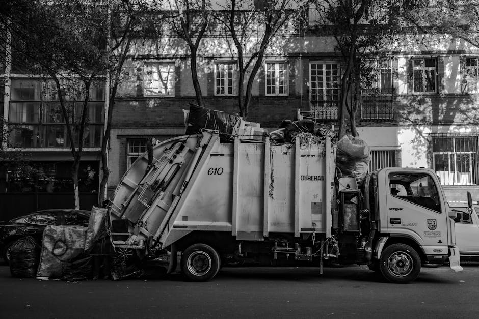 A white waste collection truck parked on an urban street in front of a multi-storey brick building with white-framed windows and black metal balcony railings. The truck's rear compartment is filled wi